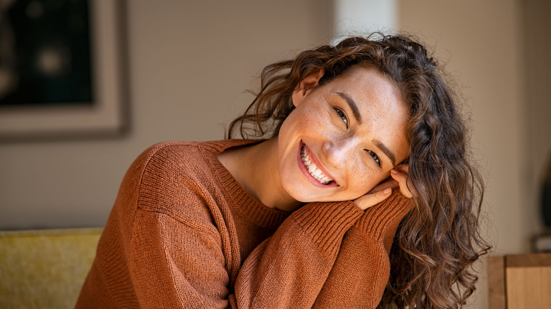Young woman with curly hair and freckles smiling warmly, resting her cheek on her hand whilst seated indoors